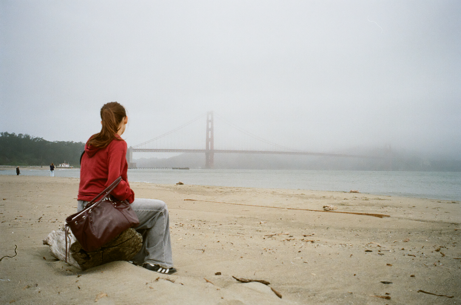 Woman seated on the beach facing a fog-covered bridge